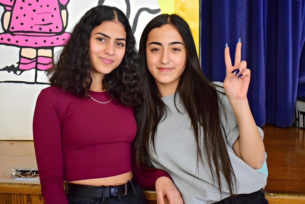 Zwei junge Frauen mit dunklen Haaren posieren vor einem farbenfrohen Wandbild auf dem Campus der Honourable W.C. Kennedy Collegiate.