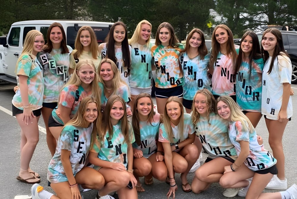 Eine Gruppe von Schülerinnen der Hopkinton High School posiert in bunten Tie-Dye-Outfits vor einem geparkten Fahrzeug im Freien.