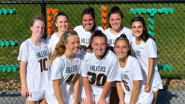 Eine Gruppe junger Sportlerinnen in weißen Uniformen posiert vor einem grünen Zaun auf dem Campus der Hopkinton High School.