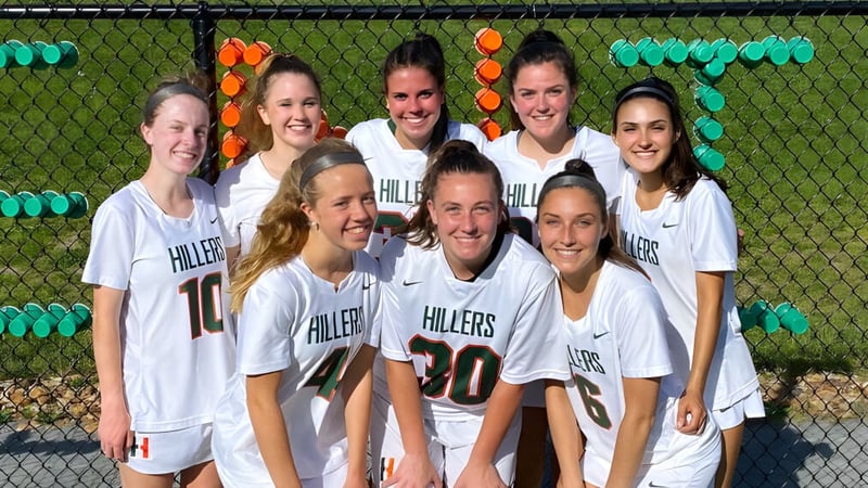 Eine Gruppe junger Sportlerinnen in weißen Uniformen posiert vor einem grünen Zaun auf dem Campus der Hopkinton High School.