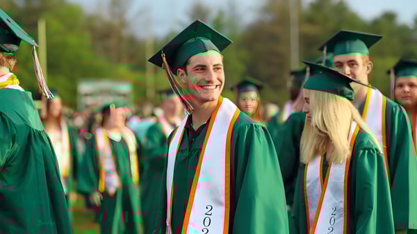 Eine Gruppe von Absolventinnen und Absolventen des Hopkinton Public School District steht in grünen und orangefarbenen Talaren vor einem Waldhintergrund.