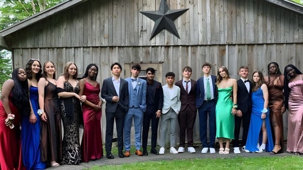 Eine Gruppe von Personen in formeller Kleidung steht vor einer rustikalen Scheune mit großem Sternemblem auf dem Campus der Houghton Academy.