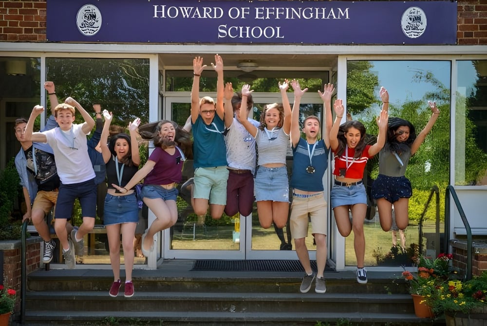 Eine Gruppe von Schülerinnen und Schülern steht mit erhobenen Armen vor der Howard of Effingham School und feiert.