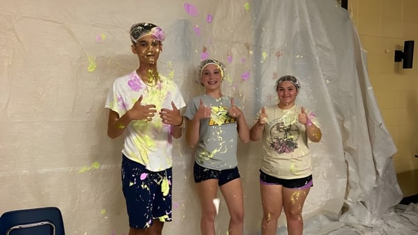 Drei Schülerinnen und Schüler der Howard S. Billings High School stehen mit bunter Farbe bedeckt in einem Raum vor einem weißen Vorhang.