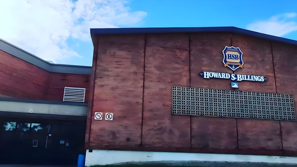 Das große Backsteingebäude der Howard S. Billings High School mit dem Logo an der Fassade vor blauem Himmel.