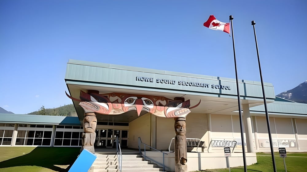 Das moderne Gebäude der Howe Sound Secondary School mit kanadischer Flagge im Hintergrund von Bergen und einer Wiesenfläche.