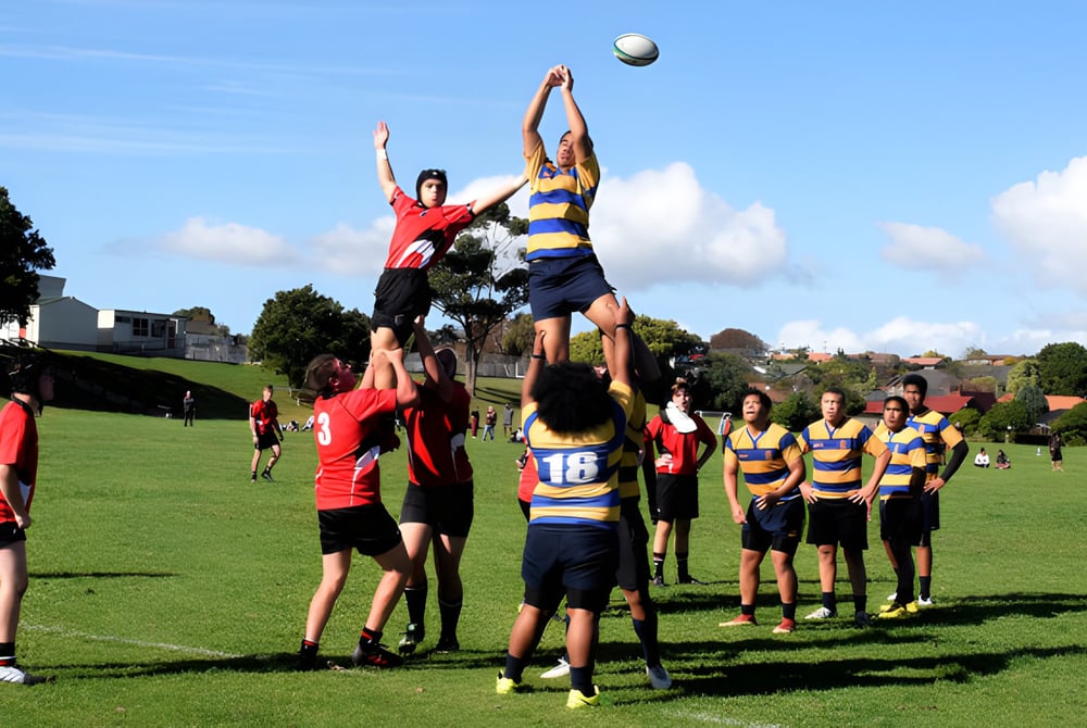 Eine Gruppe Schüler spielt Rugby auf dem Sportplatz des Howick College vor Gebäuden und Bäumen.