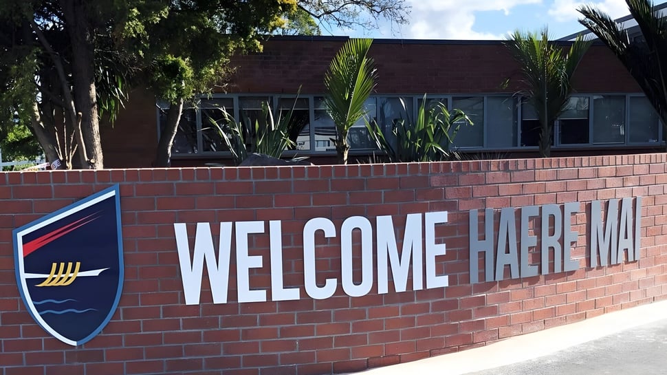 Eine Backsteinmauer mit einem großen WELCOME HAEREWA Schild auf dem Campus von Howick College, umgeben von Bäumen und Grünflächen.