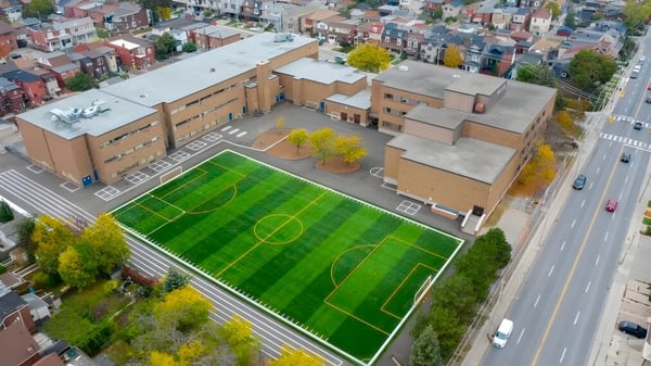 Das große grüne Fußballfeld auf dem Campus des Hudson College mit Gebäuden und einer belebten Straße im Hintergrund.