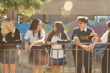 Eine Gruppe Schüler sitzt zusammen auf einer Bank im Außenbereich des Hudson College mit Bäumen und Gebäuden im Hintergrund.