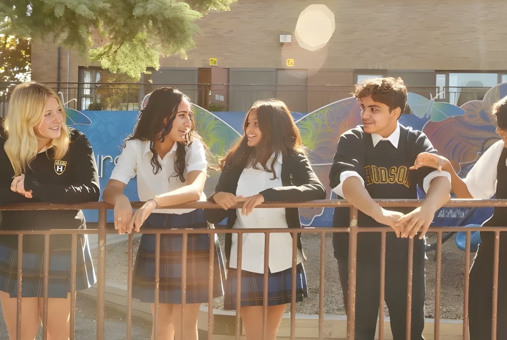 Eine Gruppe Schüler sitzt zusammen auf einer Bank im Außenbereich des Hudson College mit Bäumen und Gebäuden im Hintergrund.