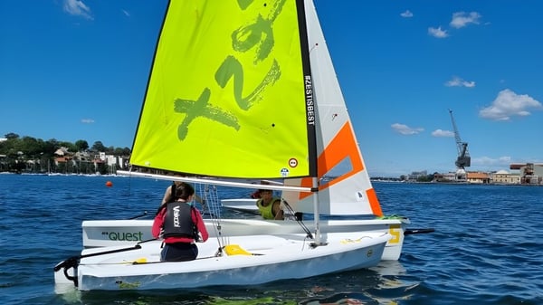 Ein kleines Segelboot mit gelb-orangefarbenem Segel segelt auf dem Wasser vor Gebäuden nahe der Hunters Hill High School.