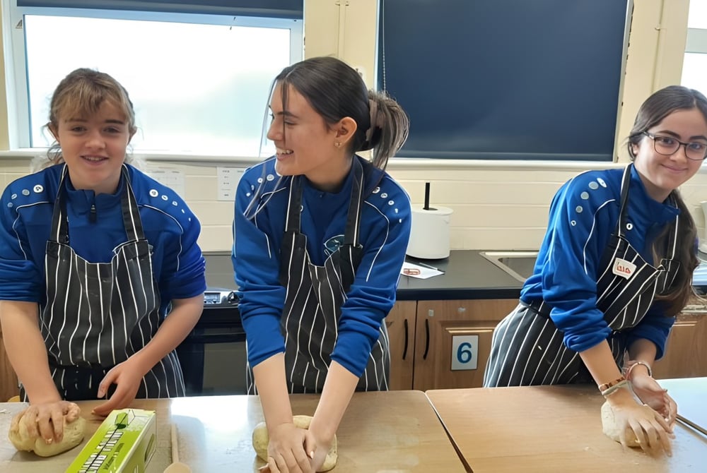 Drei Schülerinnen und Schüler des Iascaigh College stehen in blauen Uniformen an einem Tisch im Klassenraum mit Tafel im Hintergrund.