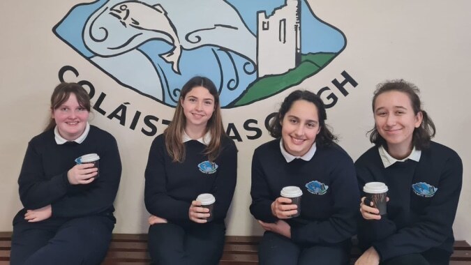 Vier Schülerinnen und Schüler in Schuluniformen stehen vor einem Wandbild mit einem schlossähnlichen Bauwerk auf dem Campus des Iascaigh College.