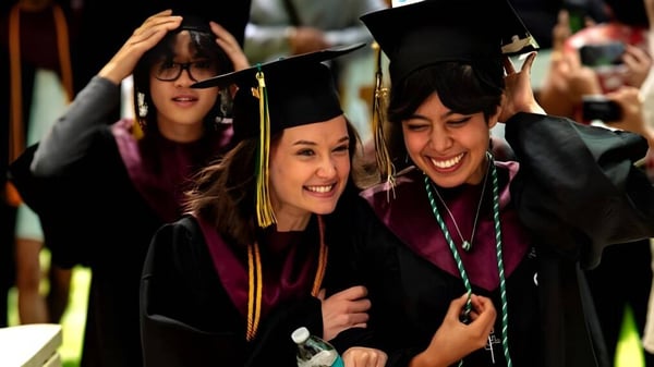 Zwei Absolventinnen der Idyllwild Arts Academy umarmen sich in ihren Abschlussroben vor einer Gruppe weiterer Graduierter.