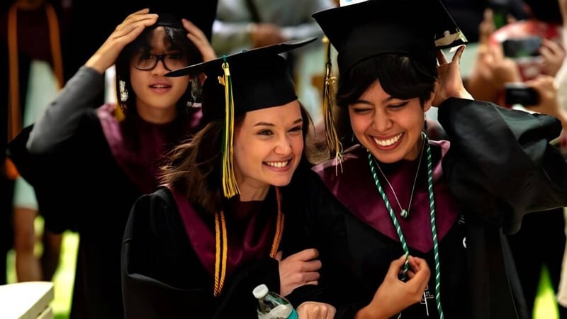 Zwei Absolventinnen der Idyllwild Arts Academy umarmen sich in ihren Abschlussroben vor einer Gruppe weiterer Graduierter.