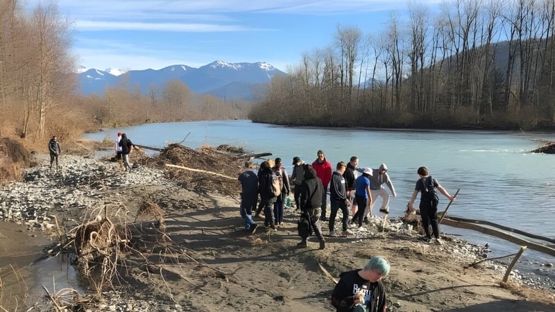 Eine Gruppe von Schülern der Imagine High Integrated Arts and Technology Secondary School geht an einem Fluss entlang auf einem matschigen Weg mit schneebedeckten Bergen im Hintergrund.