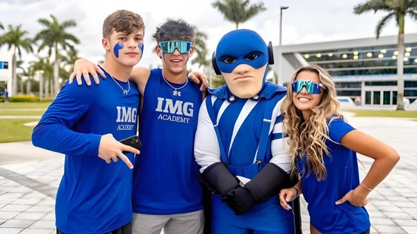 Eine Gruppe von vier Schülerinnen und Schülern trägt blaue IMG Academy Uniformen vor einem Gebäude mit Palmen.
