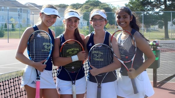 Vier Schülerinnen der Immaculata High School stehen mit Tennisschlägern auf einem Tennisplatz umgeben von Bäumen und einem Zaun.