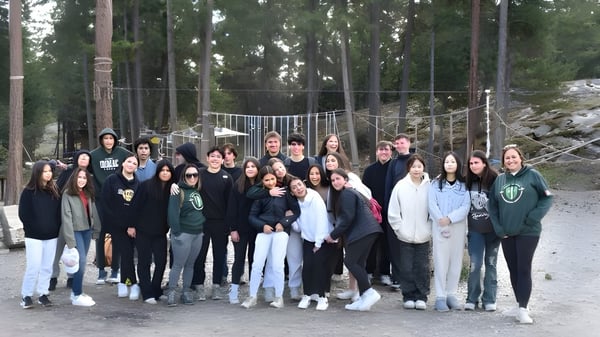 Eine Gruppe Schülerinnen und Schüler der Immaculata Regional School steht zusammen in einem bewaldeten Gebiet mit einer Seilbrücke im Hintergrund.
