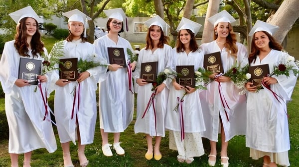 Eine Gruppe von Schülerinnen der Immaculate Heart High School steht im Freien mit ihren weißen Abschlussroben und hält ihre Diplome.