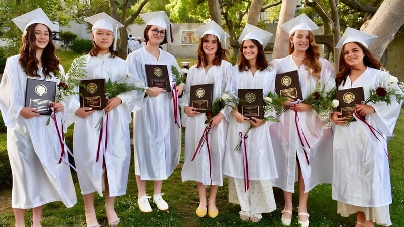 Eine Gruppe von Schülerinnen der Immaculate Heart High School steht im Freien mit ihren weißen Abschlussroben und hält ihre Diplome.