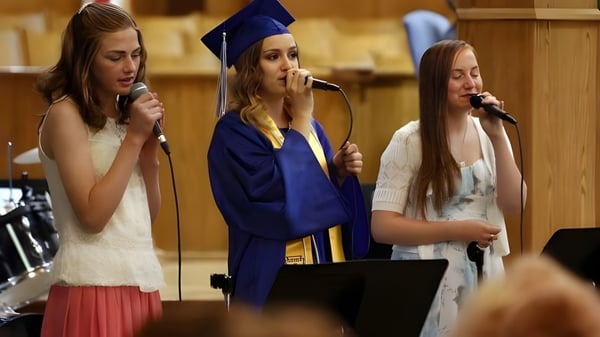 Drei Schülerinnen der Immanuel Christian Secondary School singen in Abschlussroben auf einer Bühne mit Mikrofonen.