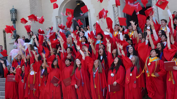 Schüler der Incarnate Word Academy versammeln sich in roten Abschlussroben auf der Treppe eines Gebäudes.