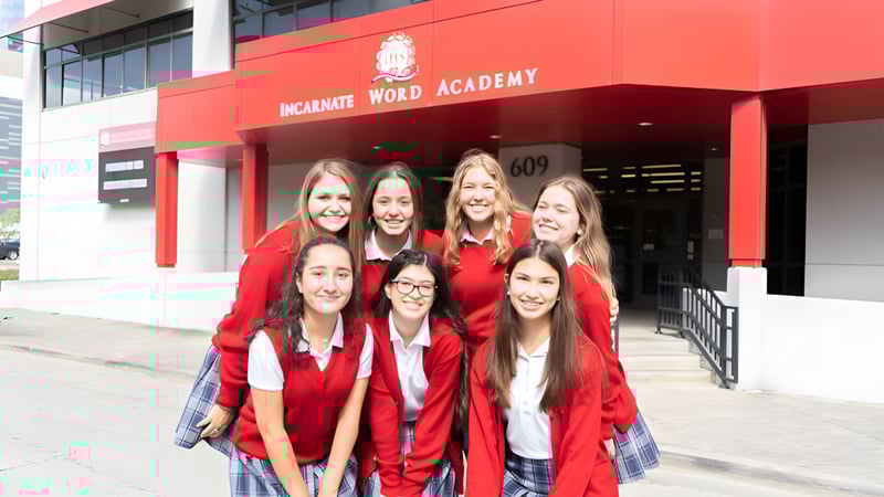 Eine Gruppe junger Frauen steht in roten Abschlusskleidern vor dem Gebäude der Incarnate Word Academy.