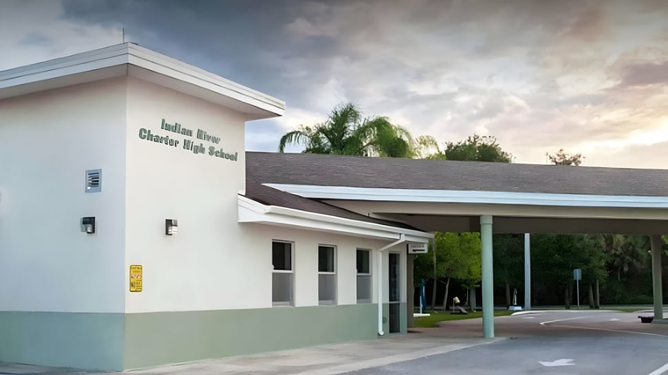 Das moderne weiße und grüne Gebäude der Indian River Charter High School mit überdachtem Eingang und Palmen im Hintergrund bei bewölktem Himmel.