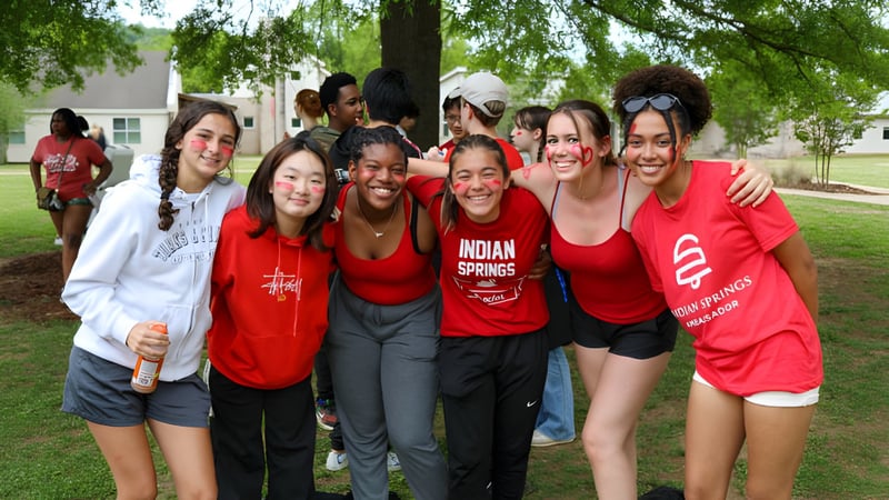 Eine Gruppe junger Frauen in roten Indian Springs School Shirts steht draußen auf einer Wiese mit Bäumen im Hintergrund.