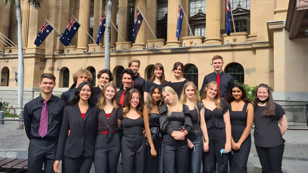 Eine Gruppe von Schülerinnen und Schülern der Indooroopilly State High School steht in formeller Kleidung vor einem großen Gebäude mit Säulen und australischen Flaggen.