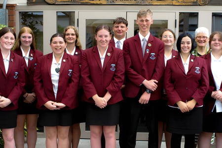 Eine Gruppe von Schülerinnen und Schülern in roten Uniformen steht vor einem Gebäude auf dem Campus der Inglewood High School mit einem Militärfahrzeug im Hintergrund.