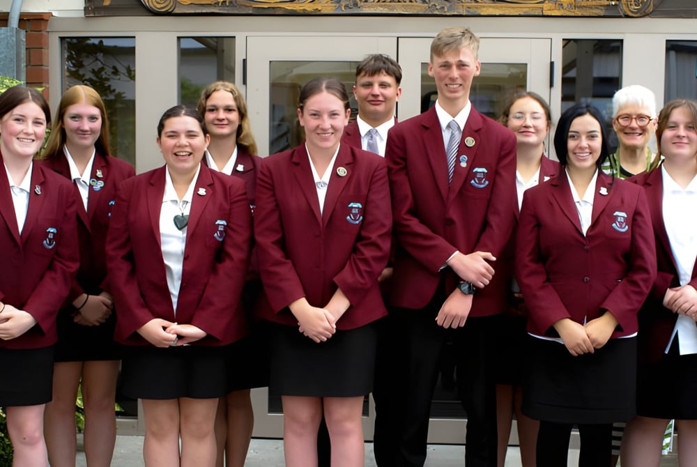 Eine Gruppe von Schülerinnen und Schülern in roten Uniformen steht vor einem Gebäude auf dem Campus der Inglewood High School mit einem Militärfahrzeug im Hintergrund.