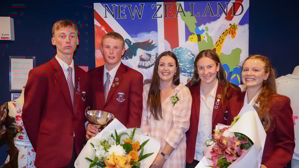 Eine Gruppe von Schülerinnen und Schülern der Inglewood High School in roten Uniformen steht mit Blumen und einem Pokal vor einer großen neuseeländischen Flagge.