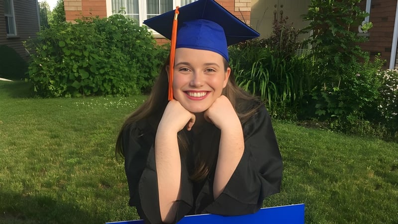 Eine lächelnde Absolventin in Talaren steht auf dem Campus der Innisdale Secondary School vor einem Backsteingebäude im Freien.