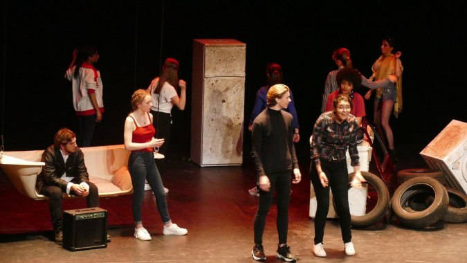 Eine Gruppe von Schauspielern steht auf der Bühne des Institut Saint-Dominique vor verschiedenen Requisiten.