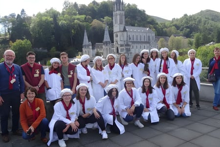 Eine Gruppe von Personen in weißen Roben steht vor dem großen verzierten Gebäude der Institution Mongazon mit Bäumen und Bergen im Hintergrund.