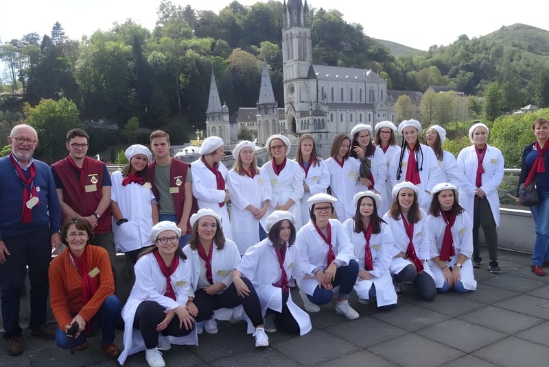 Eine Gruppe von Personen in weißen Roben steht vor dem großen verzierten Gebäude der Institution Mongazon mit Bäumen und Bergen im Hintergrund.
