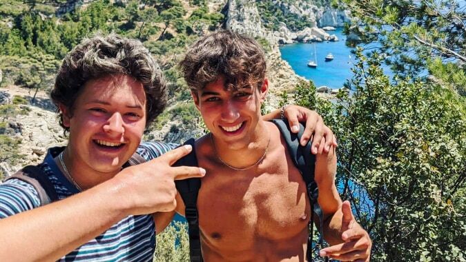 Zwei Schüler der International Bilingual School of Provence machen ein Selfie vor einer Küstenlandschaft mit Booten.