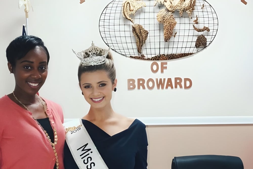 Zwei Frauen posieren vor einem Hintergrund mit dem Schriftzug der International School of Broward.