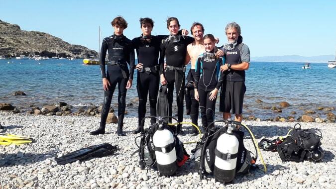 Gruppe von Schülerinnen und Schülern der International School San Patricio Toledo mit Tauchausrüstung am felsigen Strand vor Bergkulisse.