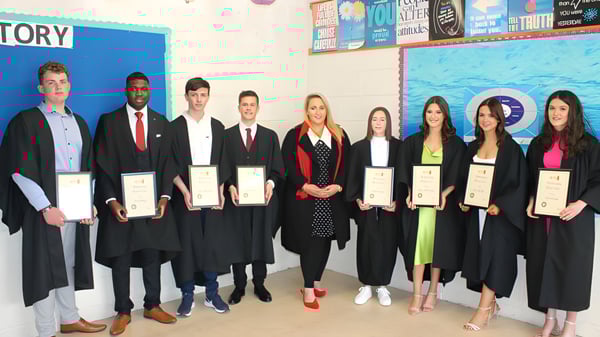 Schüler des Inver College in Abschlussroben halten ihre Zertifikate vor einer bunten Wand mit Postern.