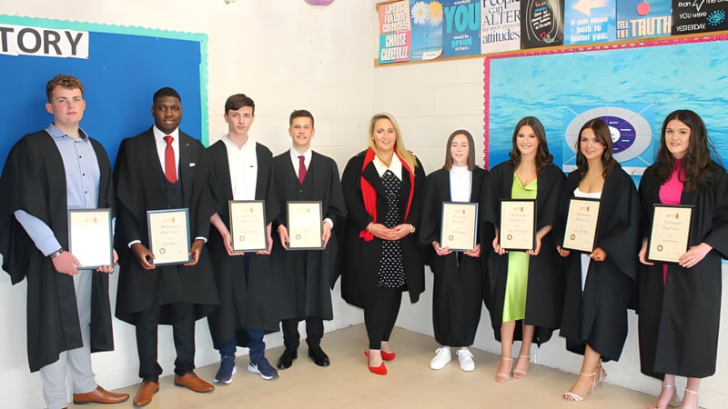 Schüler des Inver College in Abschlussroben halten ihre Zertifikate vor einer bunten Wand mit Postern.