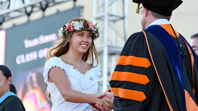 Eine Schülerin der Iolani School trägt ein weißes Kleid und einen Blumenkranz und gibt einem Mann im akademischen Gewand die Hand vor einem Universitätsgebäude.