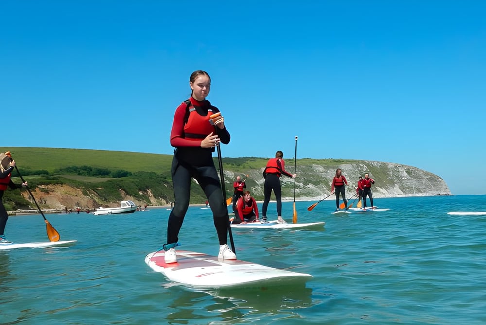 Schüler der Ipswich High School stehen auf Paddleboards in einem Gewässer mit Küstenlandschaft im Hintergrund.
