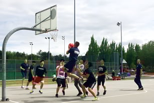 Schüler der J.N. Burnett Secondary School spielen Basketball auf einem Freiplatz unter bewölktem Himmel.