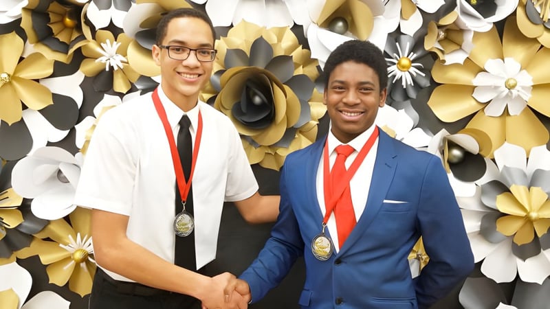 Zwei Personen schütteln sich vor einer Blumenkulisse die Hand auf dem Gelände der J. Percy Page High School.