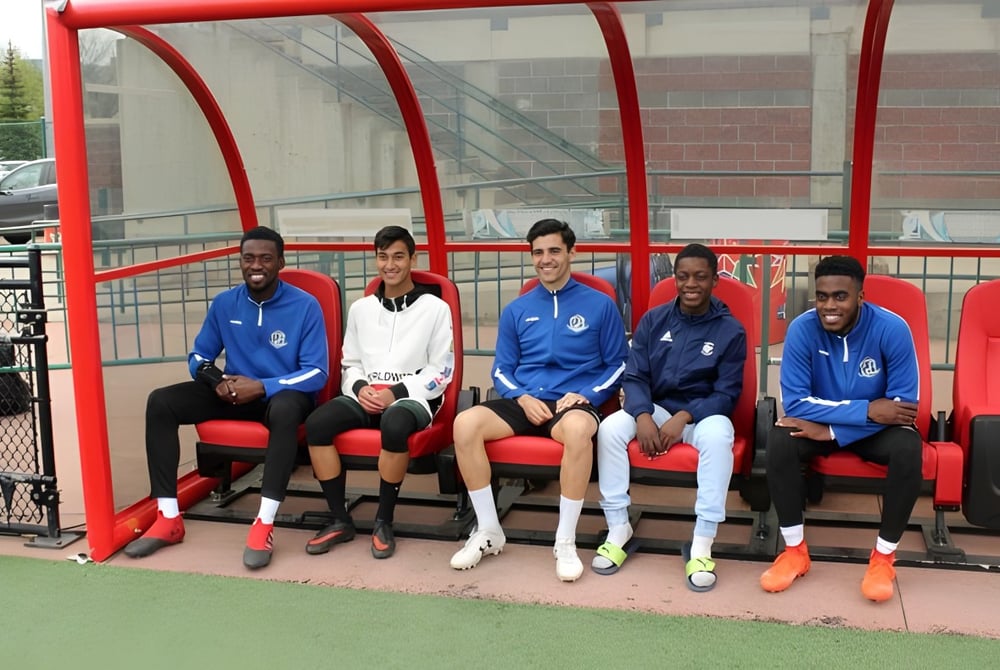 Schüler der J. Percy Page High School sitzen auf einer Bank in einer Sporthalle mit roter Struktur im Hintergrund.