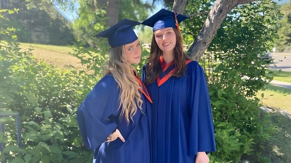 Zwei Schülerinnen der James Fowler High School stehen im Freien in ihren Abschlusskleidern mit Bäumen im Hintergrund.